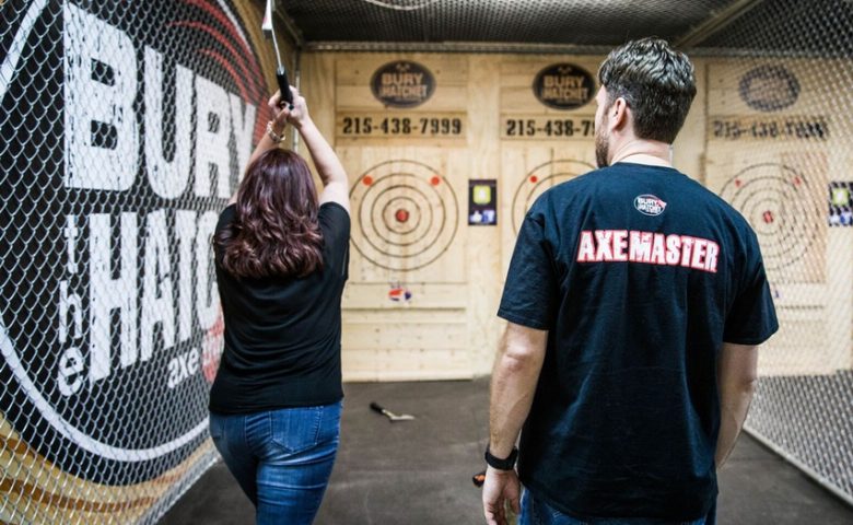 Person with axe above head facing a wooden target and another person looking on