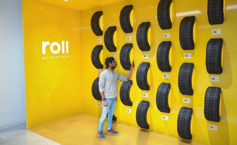 Person standing by a yellow wall that has car tires sticking out of it holding up their cell phone